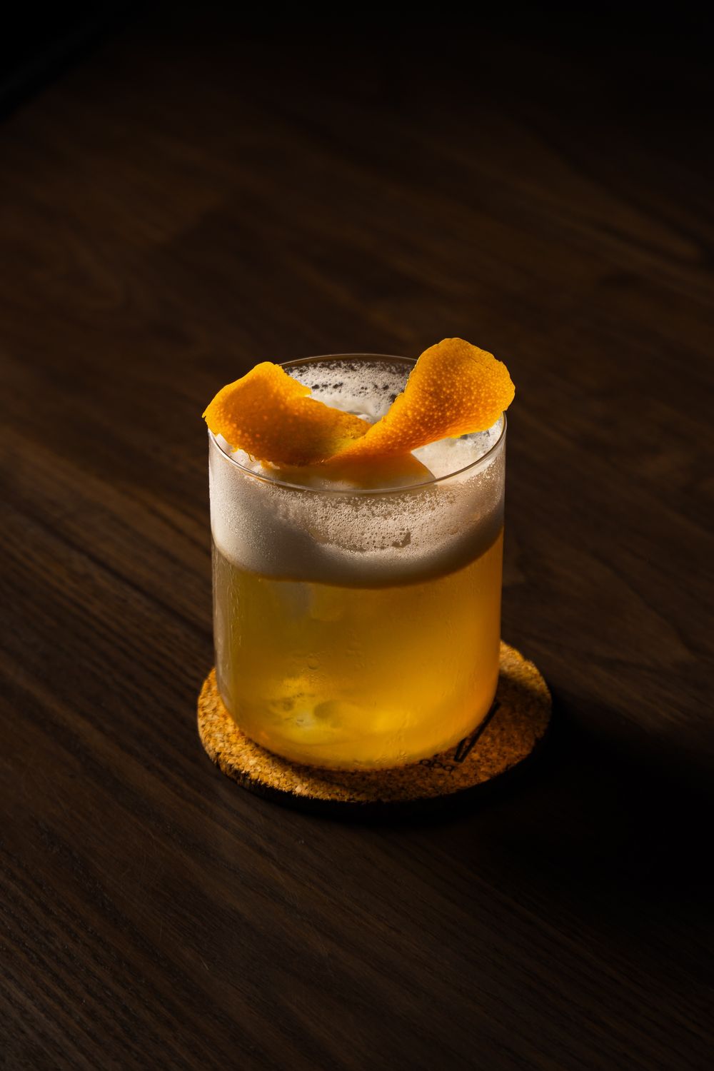 Orange-peel garnished cocktail with a foam top on a cork coaster