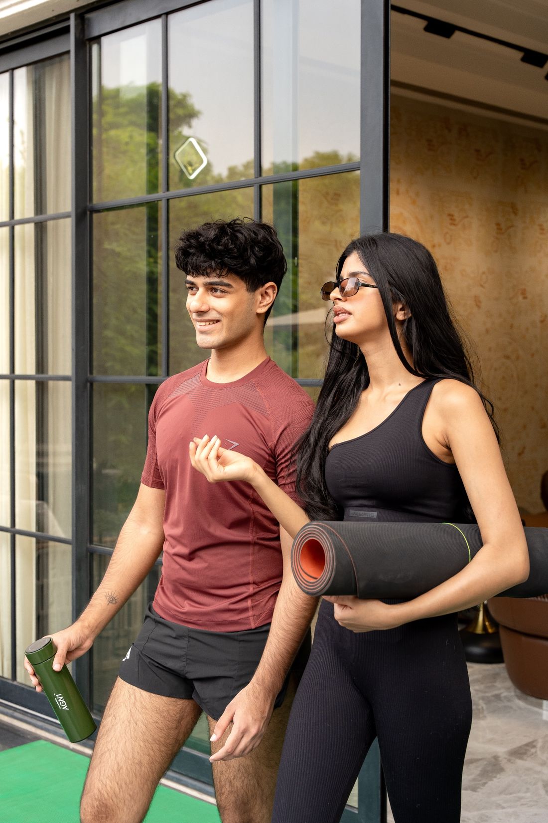 Two people in activewear on a terrace, one holding a yoga mat and the other a green bottle