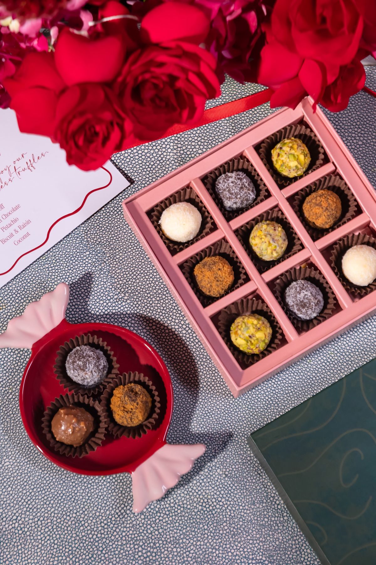 Pink box of assorted truffles beside red roses and a candy-shaped dish